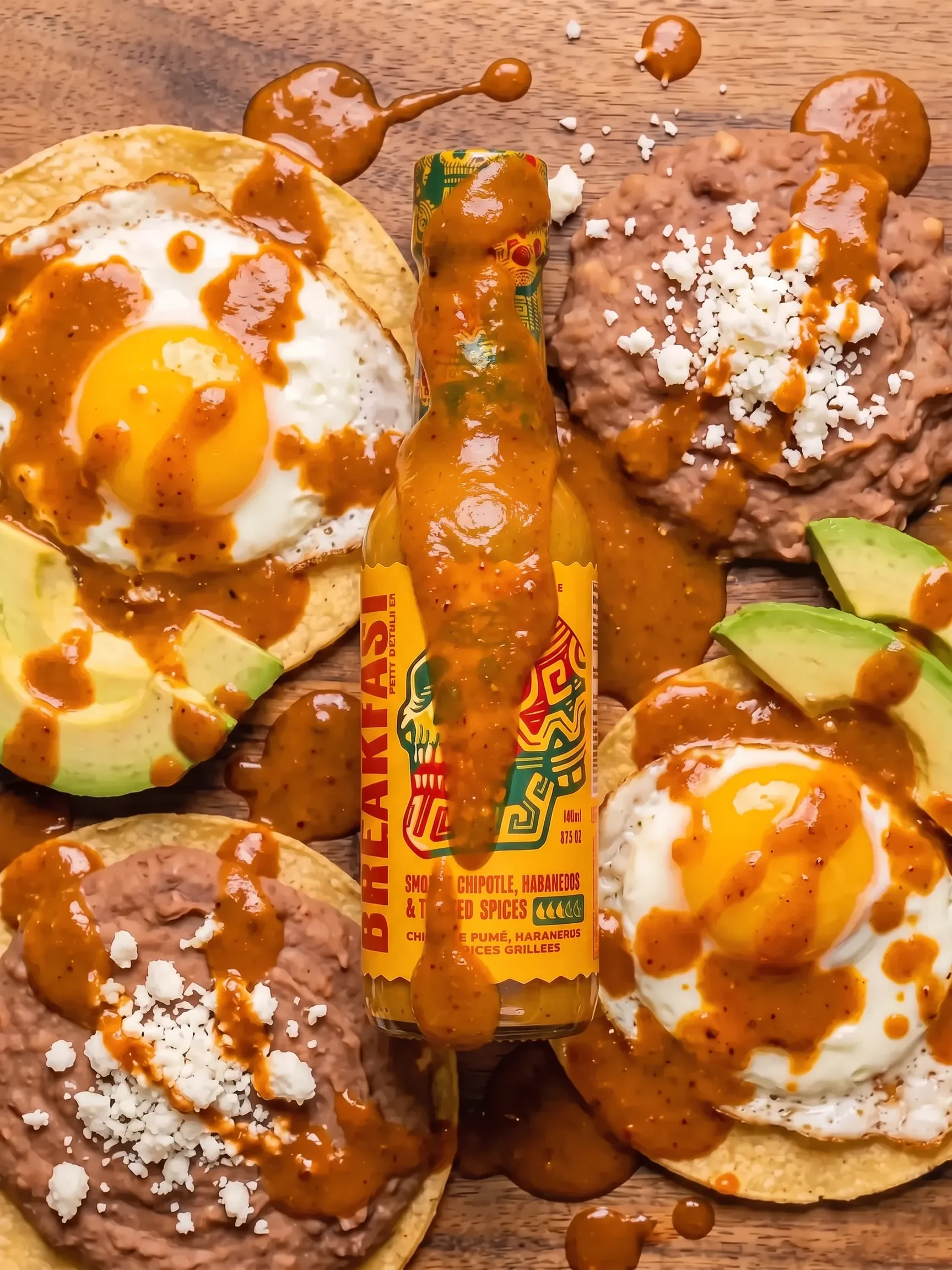 A bottle of Breakfast Spicy hot sauce centered on a wooden surface, surrounded by avocado toast, refried beans with white cheese, and eggs drizzled with orange-red sauce.